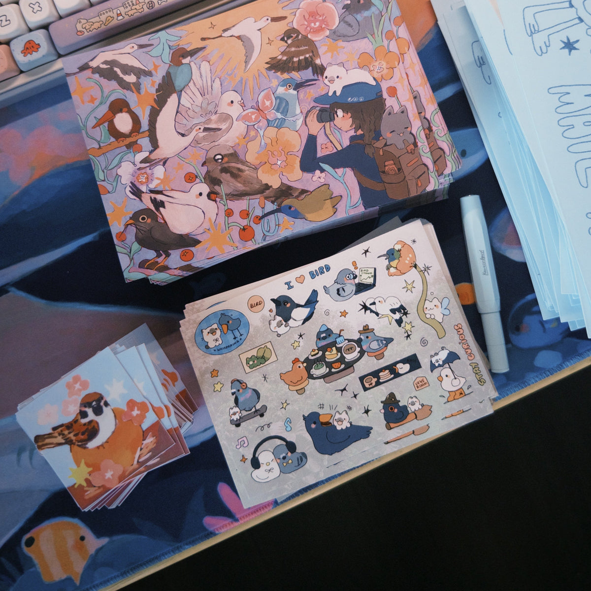 A desk setup featuring a pastel-colored mechanical keyboard, a stack of printed cards or stickers, and several sheets of white paper with blue line art and text, all arranged on a desk mat with a colorful printed surface.