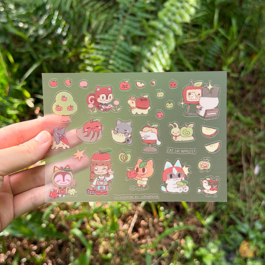 A hand holds a rectangular sticker sheet featuring cute cartoon characters and small illustrations on a muted green background, with a blurred outdoor garden setting behind it.