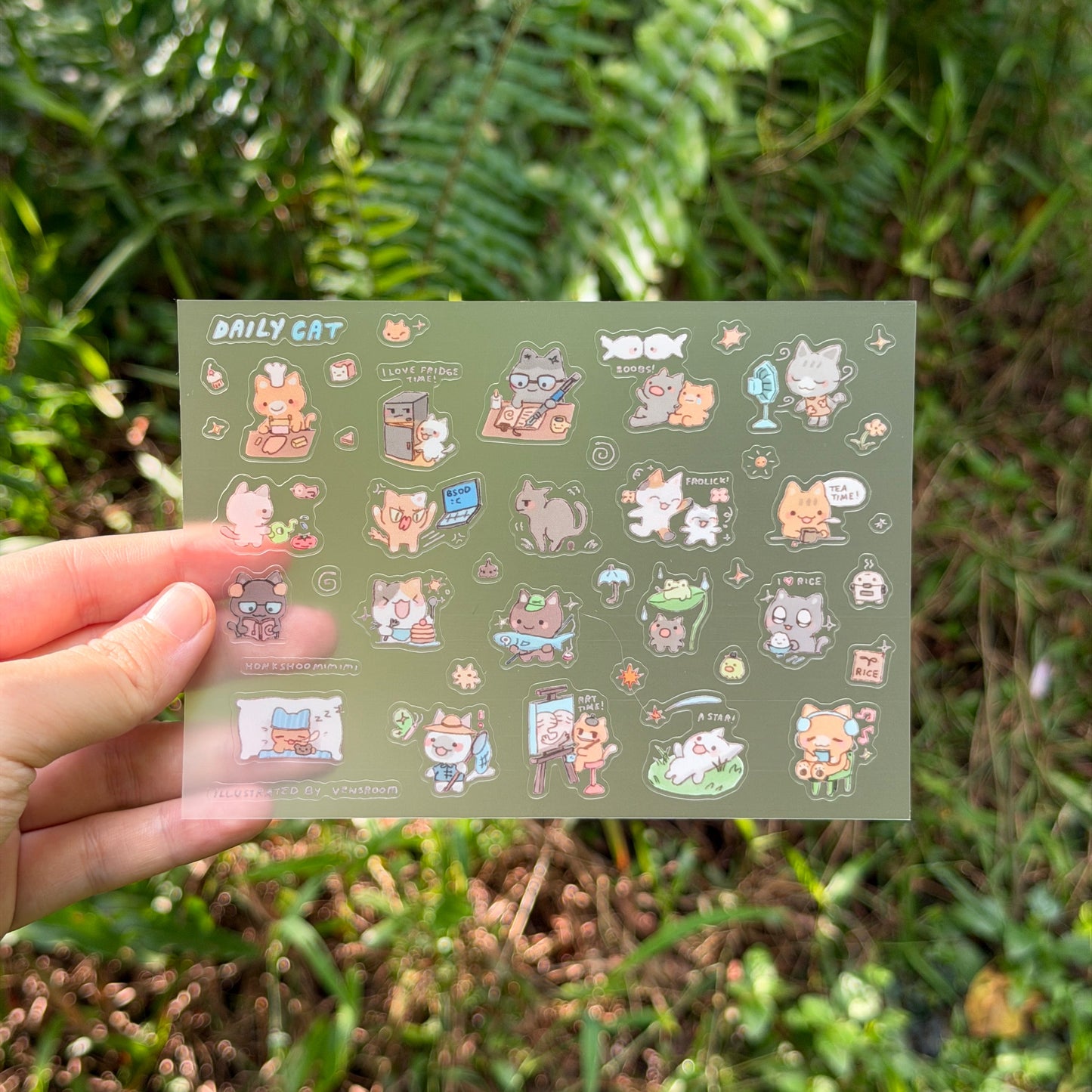 A hand holds a rectangular sticker sheet featuring cute cartoon cat illustrations against a blurred green outdoor background.