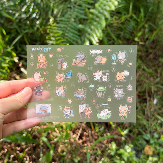 A hand holds a rectangular sticker sheet featuring cute cartoon cat illustrations against a blurred green outdoor background.