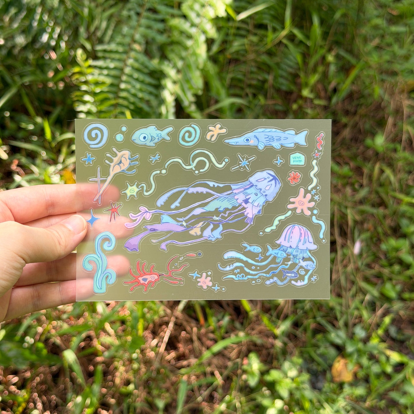 A hand holds a transparent sheet featuring colorful underwater jellyfish and sea creature illustrations against a blurred green outdoor background.
