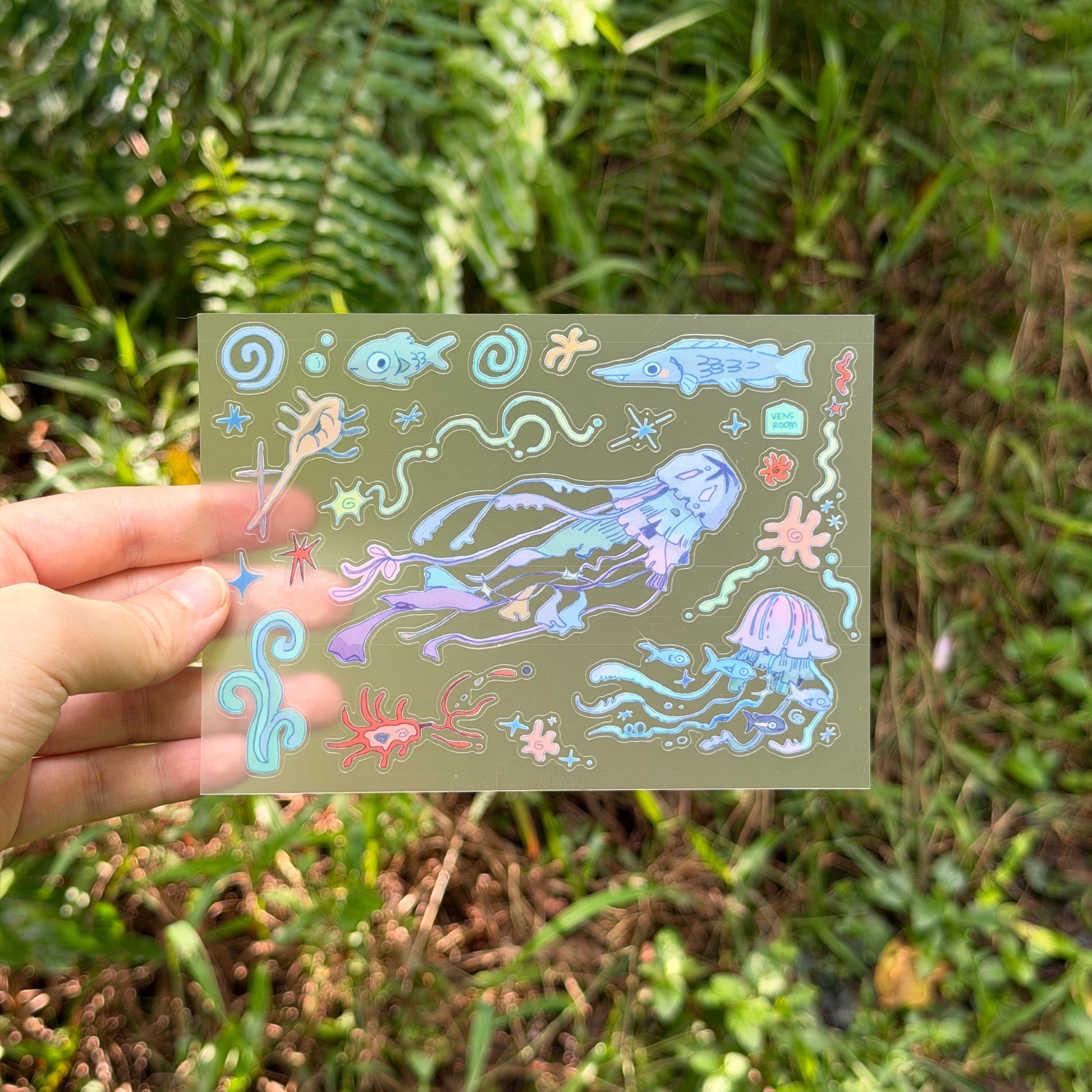 A hand holds a transparent sheet featuring colorful underwater jellyfish and sea creature illustrations against a blurred green outdoor background.