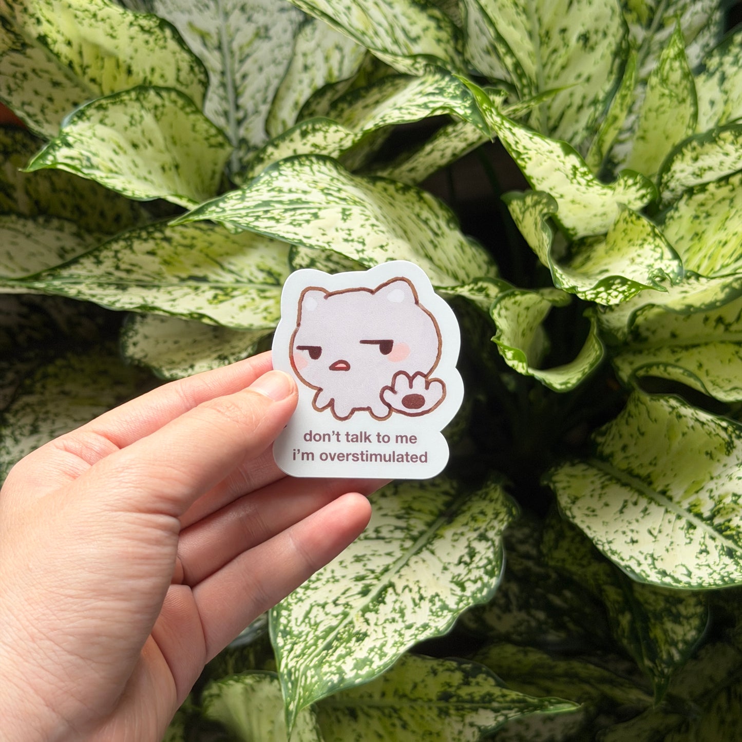 A hand holds a die-cut sticker featuring a white cartoon cat with a sad expression and the text 'don't talk to me I'm overstimulated' against a background of green and white variegated plant leaves.
