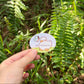A hand holding a small oval sticker featuring a cute cartoon duck and butterfly with the text 'what if it all works out' against a blurred green outdoor background.