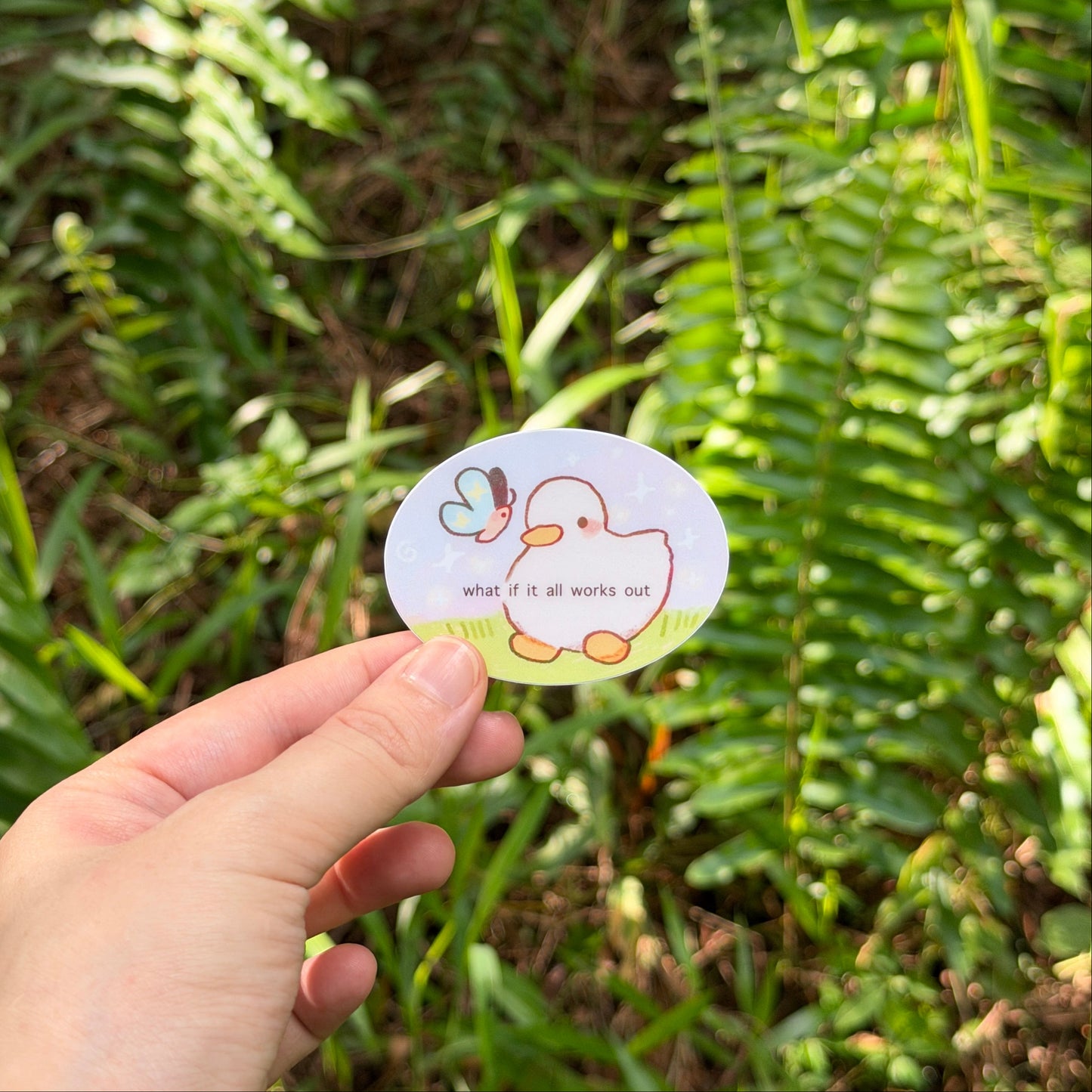 A hand holding a small oval sticker featuring a cute cartoon duck and butterfly with the text 'what if it all works out' against a blurred green outdoor background.