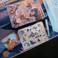 A desk setup featuring a pastel-colored mechanical keyboard, a stack of printed cards or stickers, and several sheets of white paper with blue line art and text, all arranged on a desk mat with a colorful printed surface.